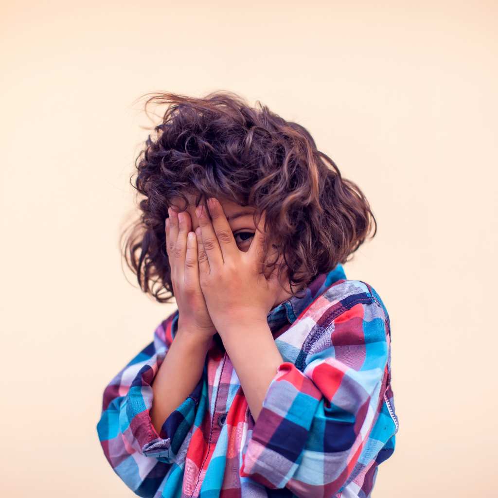 Shy child in a plaid shirt hiding their face, showing play can feel overwhelming for an autistic kid