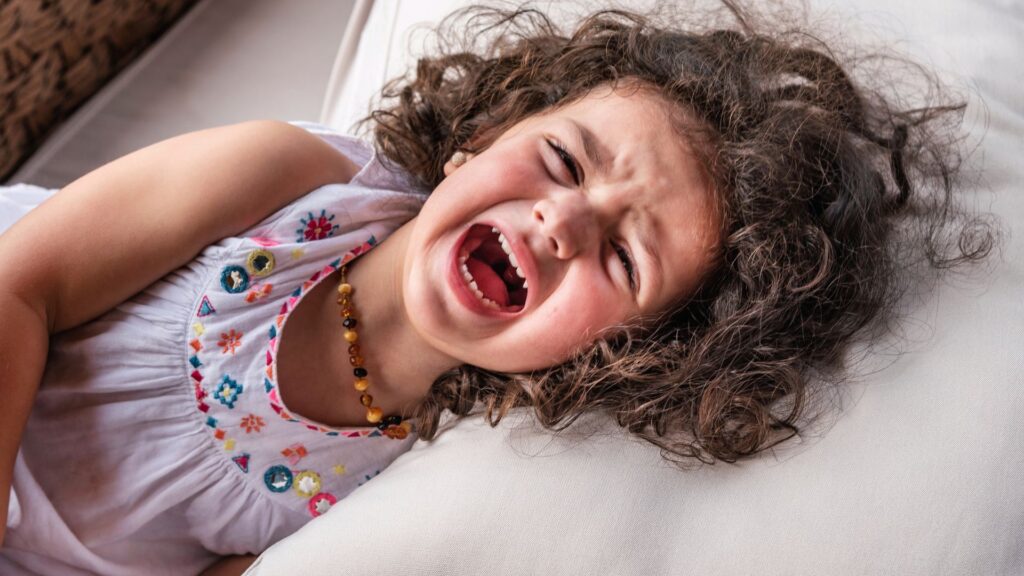 Young child experiencing toddler tantrums while lying on a couch, crying with mouth open and visibly upset during an emotional outburst.